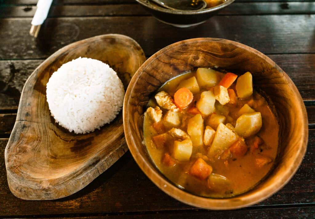 A traditional Indonesian meal served on a wooden table, featuring a mound of white rice on a wooden plate and a bowl of rich, spicy curry with chunks of chicken, potatoes, and carrots. The meal is from one of the best warungs in Canggu, Warung Ithaka