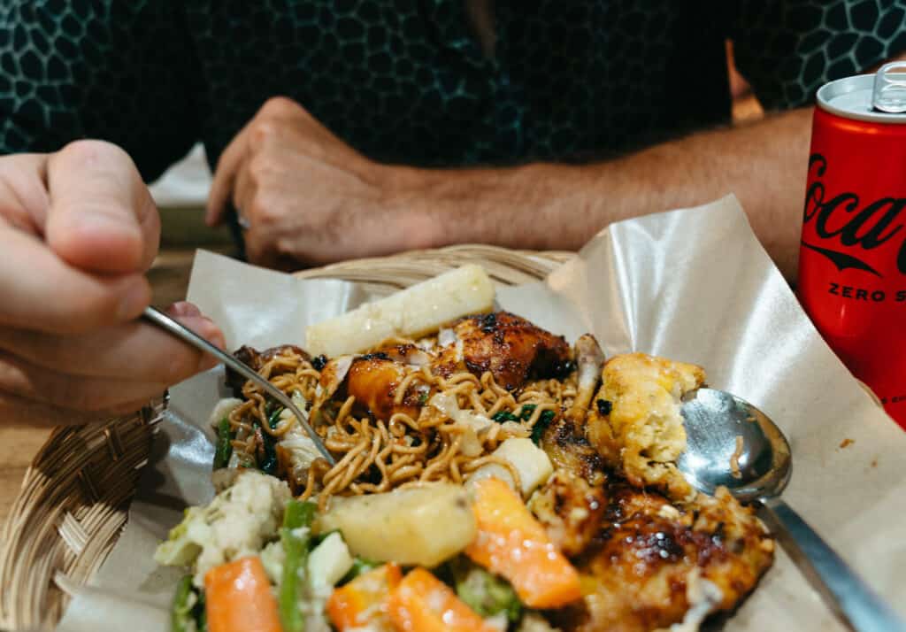 Caleb is about to enjoy a meal of grilled chicken, noodles, and assorted vegetables like carrots and cauliflower, served on a bamboo plate with a can of Coca-Cola Zero on the side. The setting appears to be casual dining, with a focus on a hearty and flavorful meal.