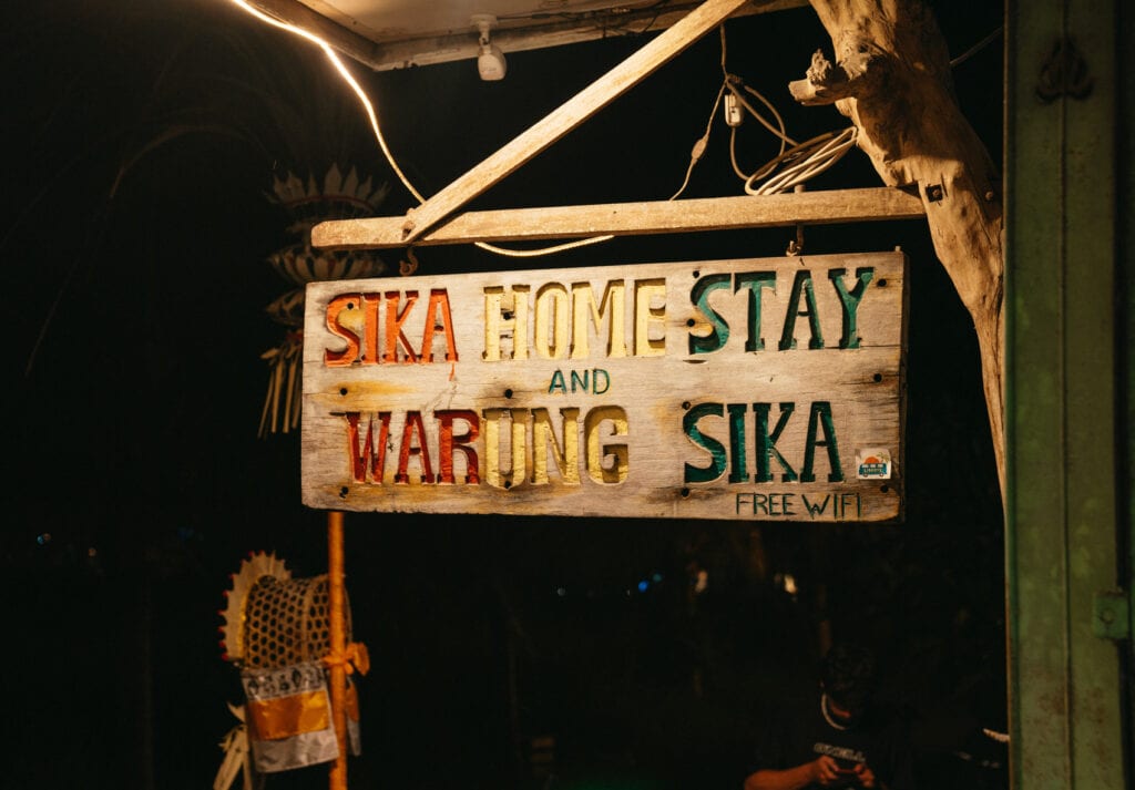 A rustic, multicolored wooden sign hangs at night, illuminated by warm light, reading 'SIKA HOME STAY and WARUNG SIKA FREE WIFI.' Below, the subtle presence of a person, indicative of the lively atmosphere typical of a Balinese homestay and eatery.