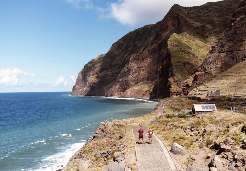 Caleb stand on a paved path along the coast, arms raised in celebration, with the dramatic cliffs and blue ocean of Madeira as their backdrop. The scene highlights the rugged beauty and exhilaration of hikes in Madeira, specifically along the Achadas da Cruz trail.