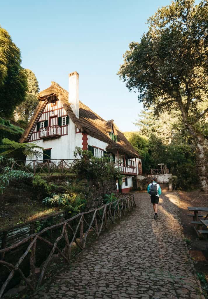 Caleb walks along a cobblestone path toward a charming, traditional house surrounded by lush greenery and large trees at the start of Caldeirão Verde hike in Madeira, Portugal.
