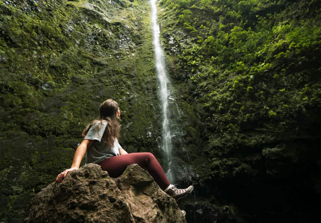 Sarah sits on a large rock, smiling and looking up at a tall, narrow waterfall at Caldeirão Verde in Madeira, Portugal.
