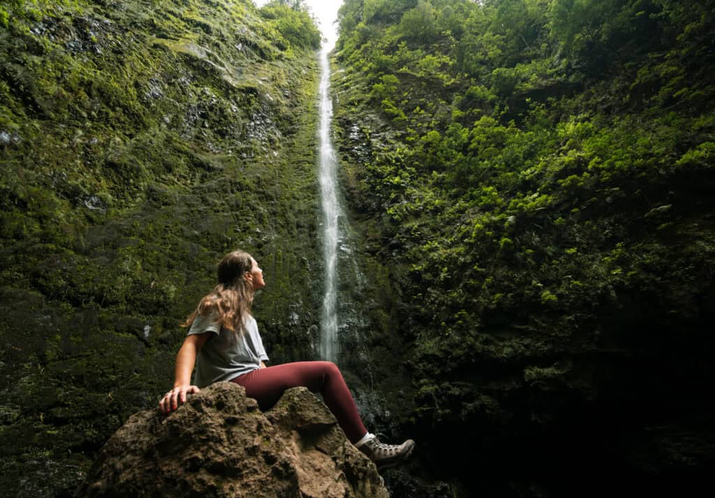 Sarah looking up at Caldeirao Verde Waterfall amidst a deep green gorge. She's wearing purple leggings and a gray T-shirt.