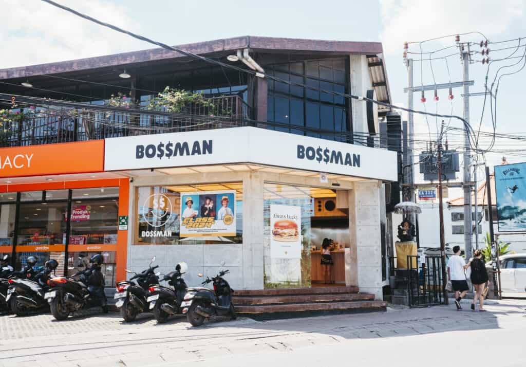 A street view of Bo$$man, a modern fast-food restaurant with a white and orange exterior. The storefront features large windows displaying advertisements, including one for a burger and another promoting a special offer.