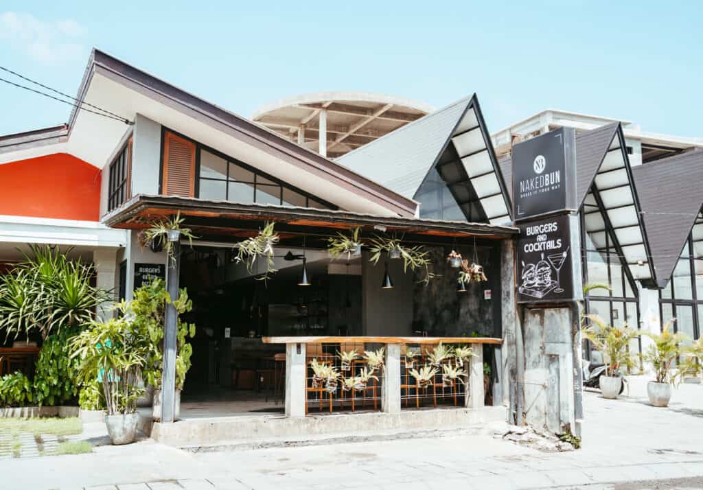 A street view of Naked Bun, a modern restaurant with an open-air design and a sleek black sign featuring a cocktail glass and the text "BURGERS AND COCKTAILS." The exterior includes a small bar area with hanging plants and a mix of contemporary and rustic elements.