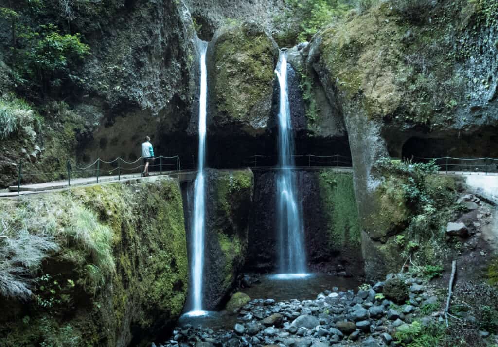 Caleb stands on a narrow bridge at Levada Nova, admiring a twin waterfall cascading down moss-covered cliffs into a rocky pool below. The scene is set in a lush, verdant gorge, capturing the serene and picturesque nature of hikes in Madeira.
