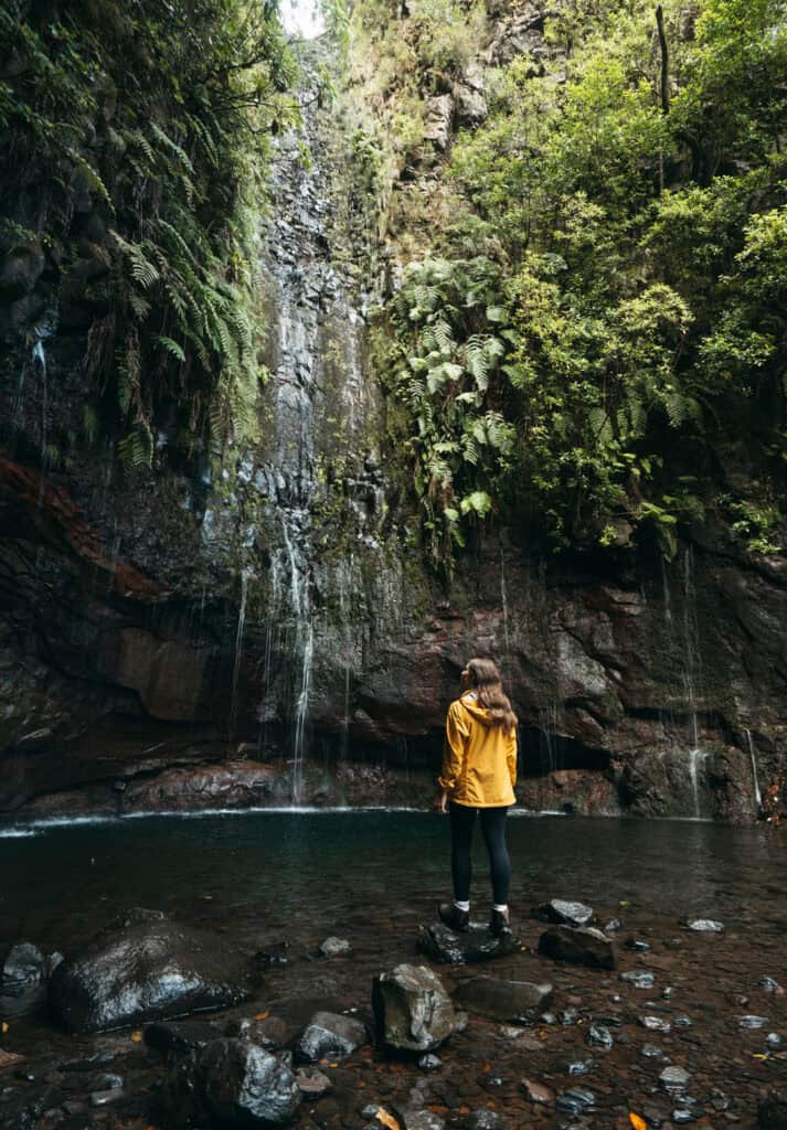 Sarah stands on rocks, gazing at a small waterfall cascading into a clear pool surrounded by lush greenery. The scene is set in the Levadas 25 Fontes in Madeira, Portugal, with tall cliffs covered in moss and ferns framing the waterfall.
