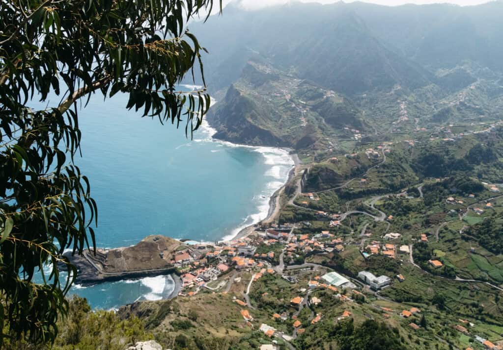 Aerial view of a coastal village nestled in a valley, with houses and winding roads stretching along the shoreline and up into the green hills. The scene is framed by tree branches in the foreground, capturing the scenic beauty of Penha de Águia in Madeira, Portugal.
