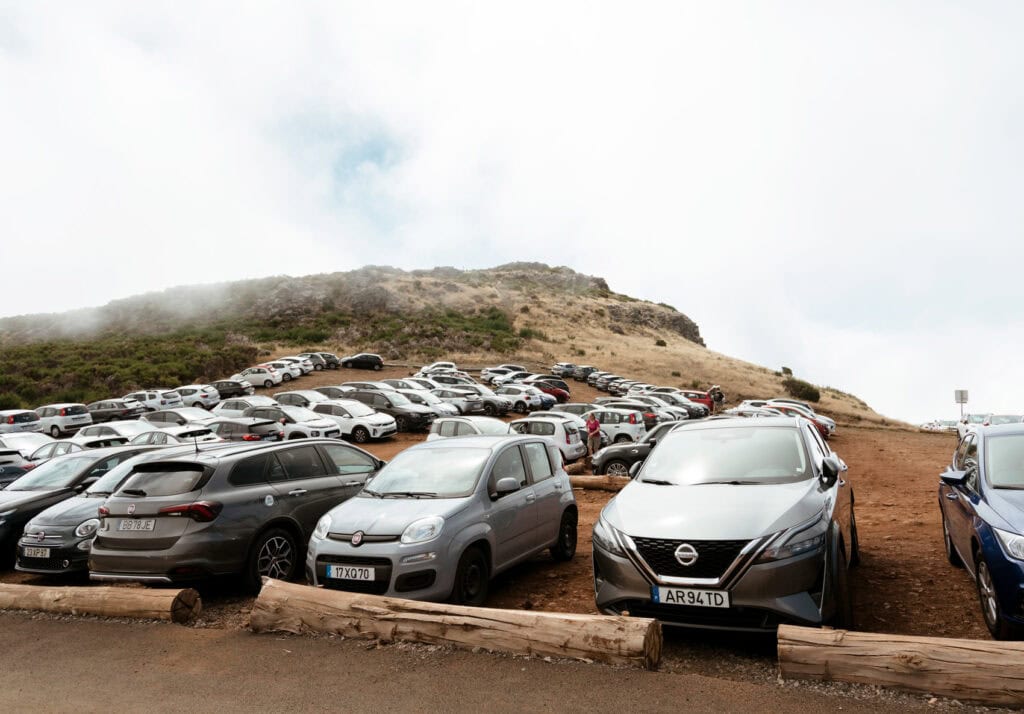 A large parking lot filled with cars, set against a backdrop of rocky hills at the base of Pico do Arieiro in Madeira, Portugal.