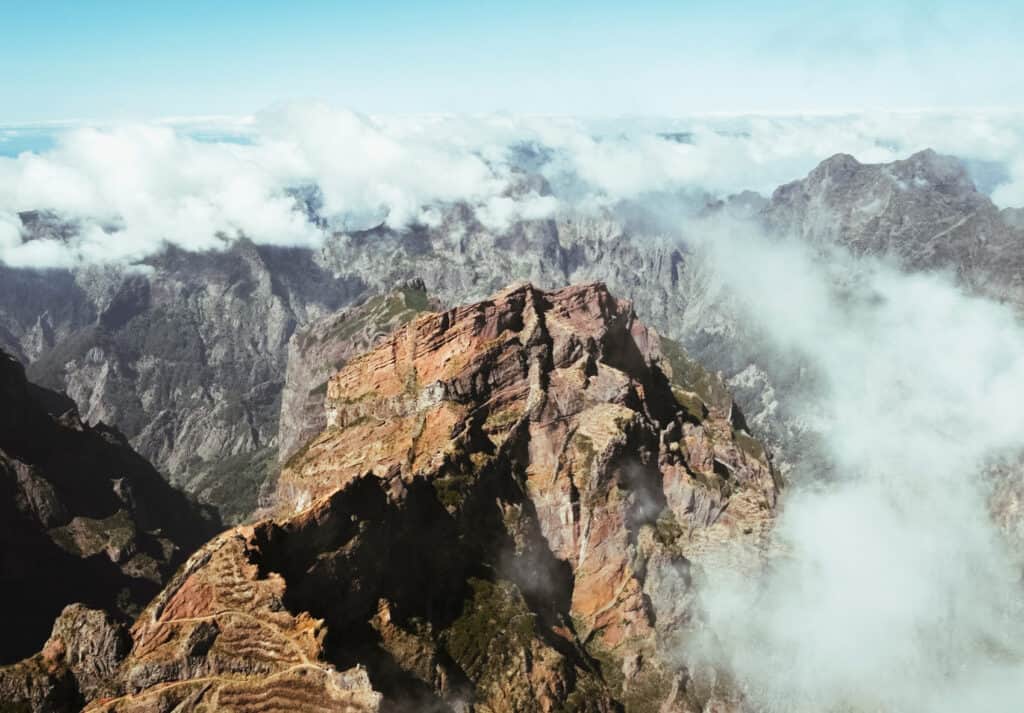Aerial view of Pico do Areerio mountain in Madeira, Portugal, surrounded by clouds. The rugged, rocky peaks are prominent, with layers of reddish and brown hues, contrasting against the green valleys below.