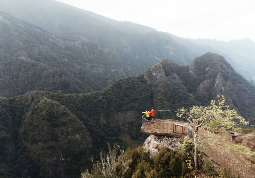 Caleb and Sarah stand on a lookout platform with a metal railing, gazing out at the lush, green mountains and valleys of Madeira. The platform juts out over the landscape, providing a stunning panoramic view of the surrounding natural beauty from the Vereda dos Balcões trail.