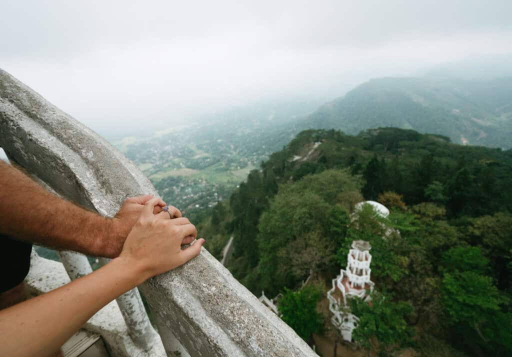 holding hands on the railing of the Ambuluwawa Tower, overlooking misty green hills and valleys below. This scenic viewpoint offers breathtaking views of the surrounding landscape from the heights of the tower in Sri Lanka.