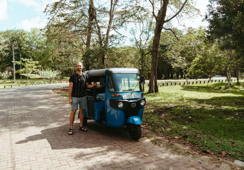 Caleb stands next to a blue tuk-tuk on a tree-lined path in Anuradhapura, Sri Lanka. Caleb and Sarah are exploring the historic city by tuk-tuk.