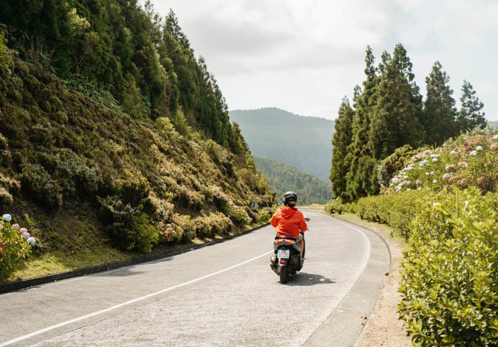 Caleb rides a scooter down a scenic road in the Azores, surrounded by lush greenery and vibrant flowers. The road curves ahead, framed by tall trees and mountains in the background.