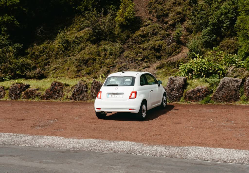 White car parked on a dirt parking area surrounded by greenery and rocks, located near a hillside in a rural area outside Ponta Delgada.