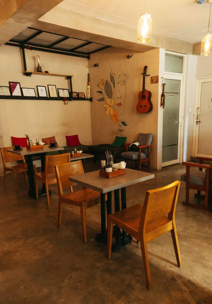 The interior of a cozy café with minimalistic decor. The space is furnished with simple wooden tables and chairs, with a few colorful cushions on a bench along the wall. The lighting is warm, creating a welcoming atmosphere.