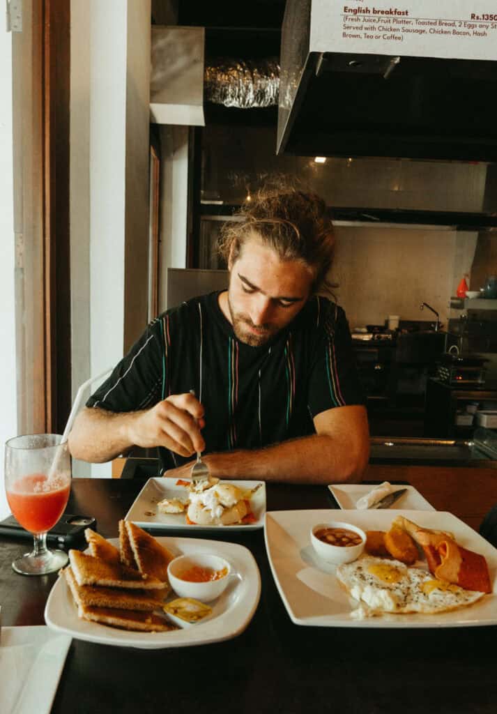 Caleb enjoying breakfast at a café. The table has a plate of eggs Benedict and plates of toast with jam, fried eggs, baked beans, and hash browns.