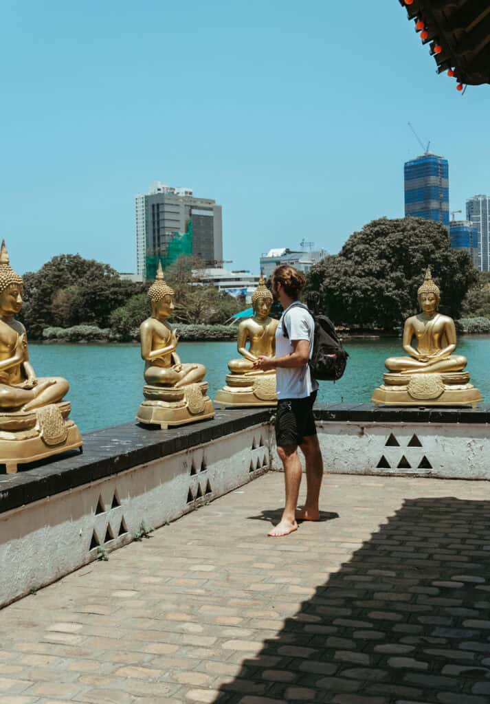 Caleb standing barefoot in front of golden Buddha statues at Gangaramaya Temple in Colombo, Sri Lanka, with a lake and modern skyscrapers visible in the background, blending tradition with urban development.