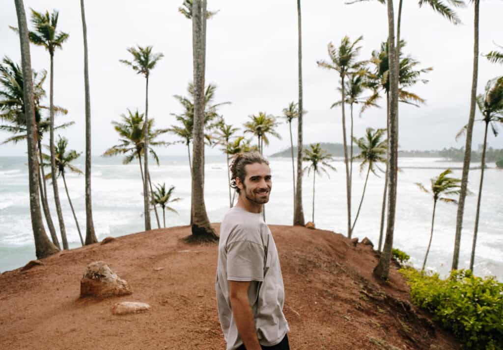 Caleb standing on Coconut Hill in Mirissa, Sri Lanka, surrounded by tall palm trees overlooking a scenic ocean view with waves crashing onto the shore in the background.