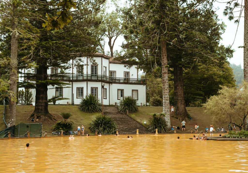 People swimming in the yellow-brown thermal pool at Parque Terra Nostra in the Azores, with a white building nestled among tall trees in the background.