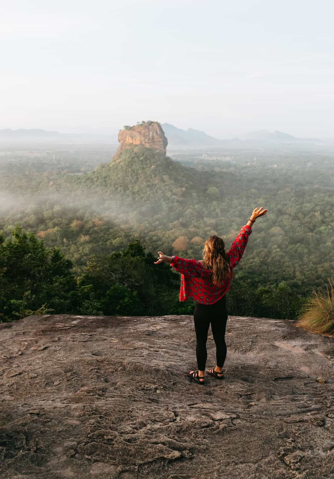 Sarah stands on a rocky ledge at Pidurangala Rock in Sri Lanka, as she gazes at the distant Sigiriya Rock Fortress from across the way.
