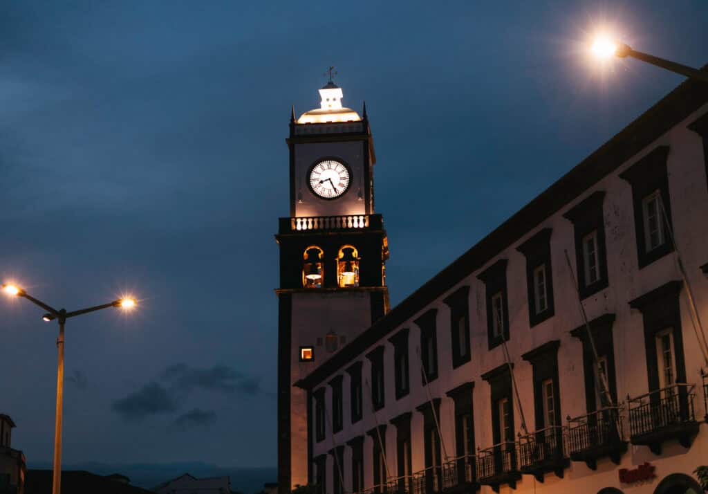 Nighttime view of a clock tower in Ponta Delgada, brightly lit against a darkening sky, with streetlights illuminating the scene. The adjacent building showcases traditional architecture.