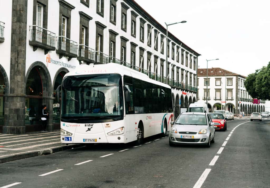 Tour bus parked alongside a busy street in Ponta Delgada, with cars passing by in front of a historic building featuring arched windows and balconies.