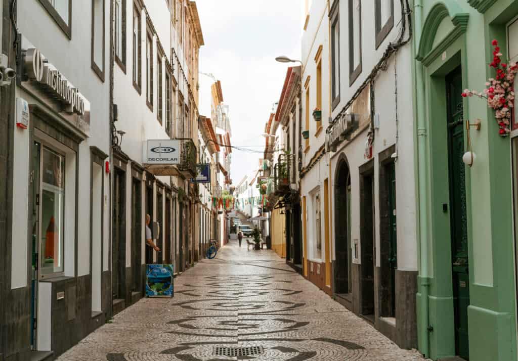 Narrow cobblestone street in Ponta Delgada, lined with colorful buildings, small shops, and balconies adorned with flowers. The street is quiet, with a few people walking in the distance.