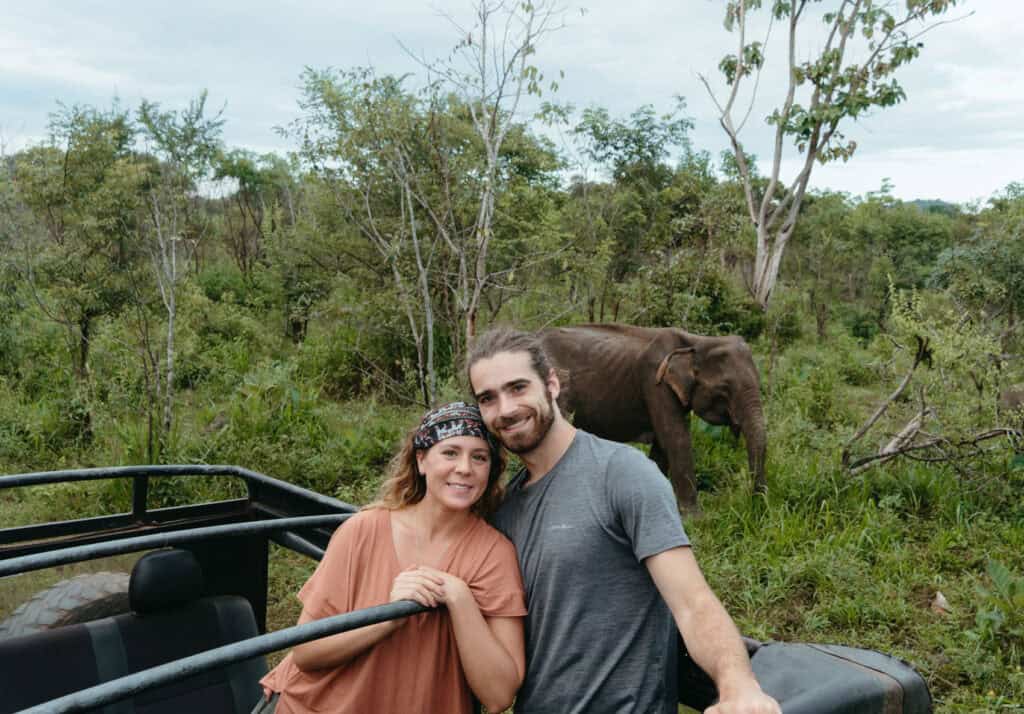 Caleb and Sarah smile as they are leaning on a safari vehicle during a one-day trip from Colombo, with a wild elephant grazing in the background while exploring Sri Lanka's wildlife on safari.