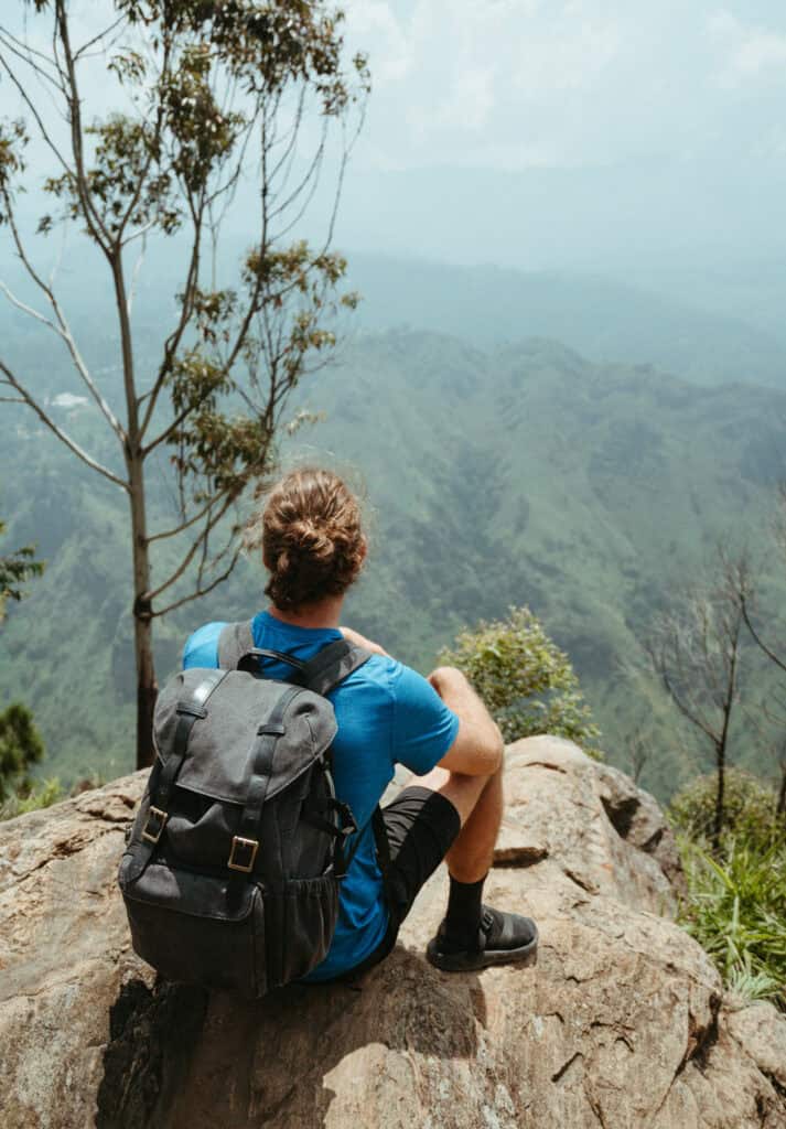 Caleb sitting on a rocky ledge at Ella Rock in Sri Lanka, gazing out at a panoramic view of lush green mountains and valleys.