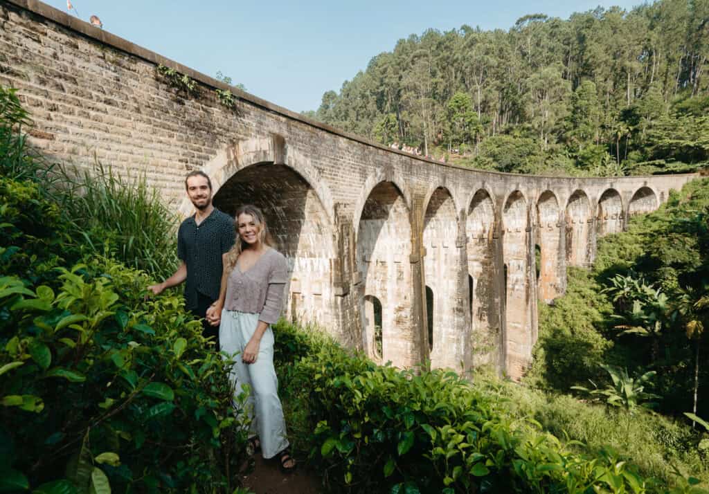 Caleb and Sarah standing near the iconic Nine Arches Bridge in Ella, Sri Lanka., with the scenic stone arches and a backdrop of dense forest.