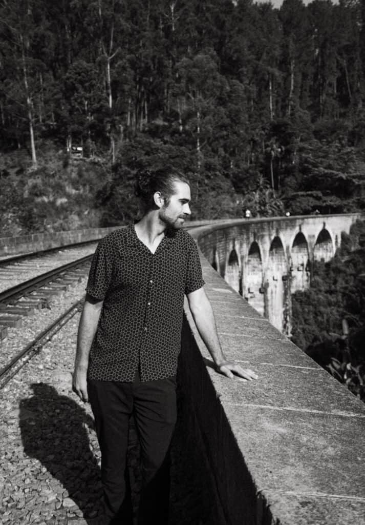 Black and white portrait of Caleb standing on the Nine Arches Bridge in Ella, Sri Lanka, leaning on the stone railing.