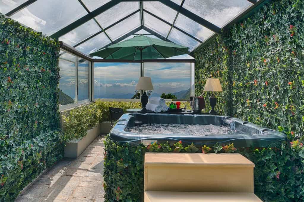 A luxurious indoor hot tub at EKHO Ella Hotel enclosed by a glass roof and walls adorned with lush green ivy. The space is styled with elegant decor, including lamps and an umbrella, and offers a stunning view of Ella Rock and clouds in the distance.