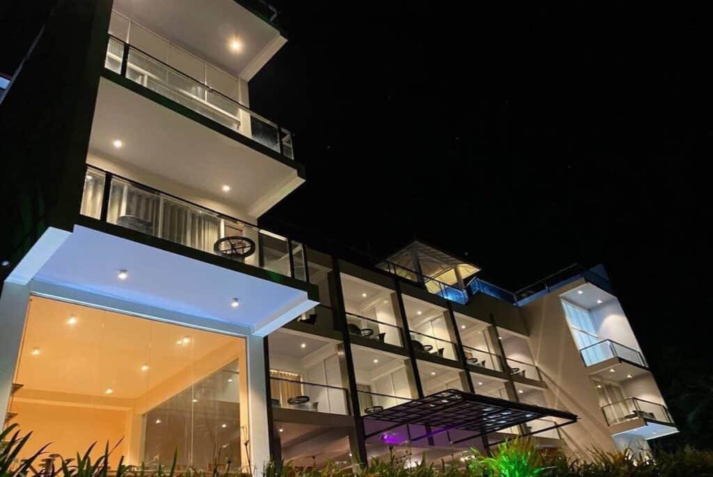 A modern multi-story building illuminated at night, featuring large glass windows, open balconies with seating, and contemporary architectural design surrounded by greenery.