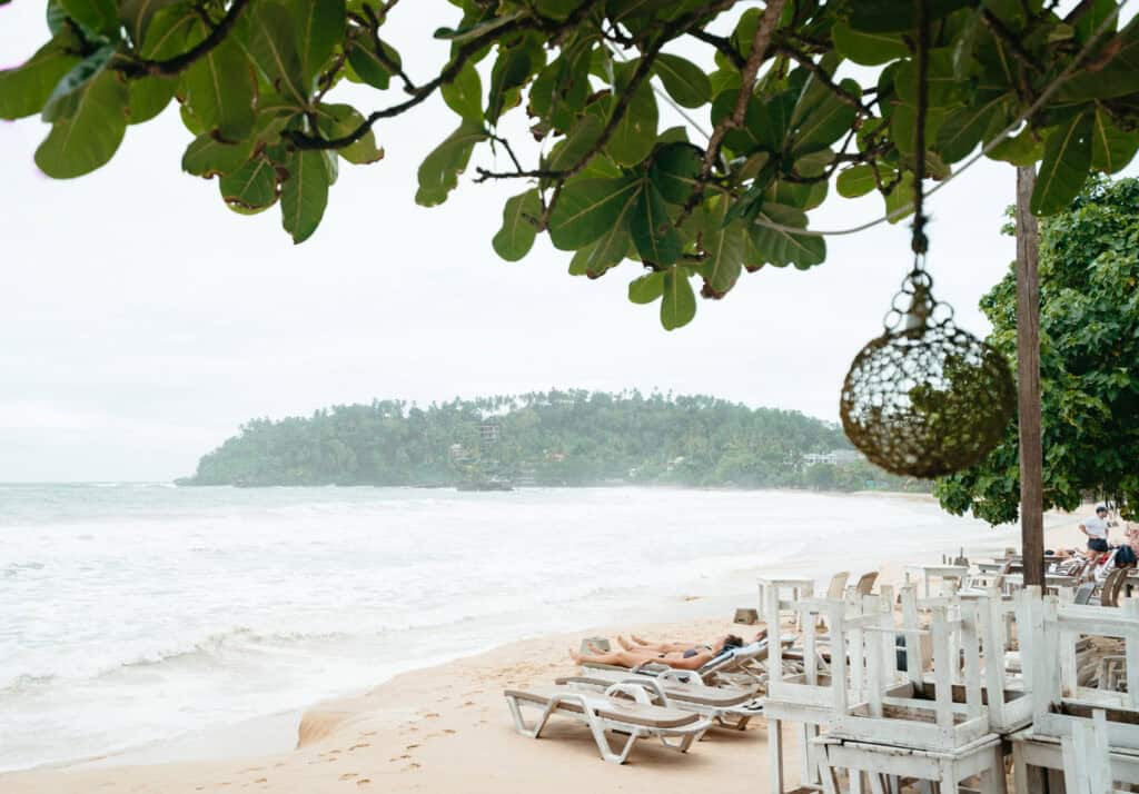 A serene view of Mirissa Beach in Sri Lanka, framed by green foliage and a decorative hanging ornament, with waves crashing onto shore