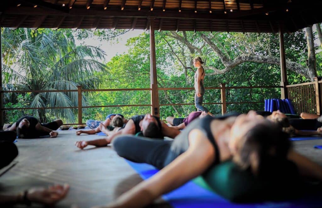 A serene outdoor yoga session under a wooden pavilion, with participants lying on mats surrounded by lush green trees and a tranquil atmosphere.