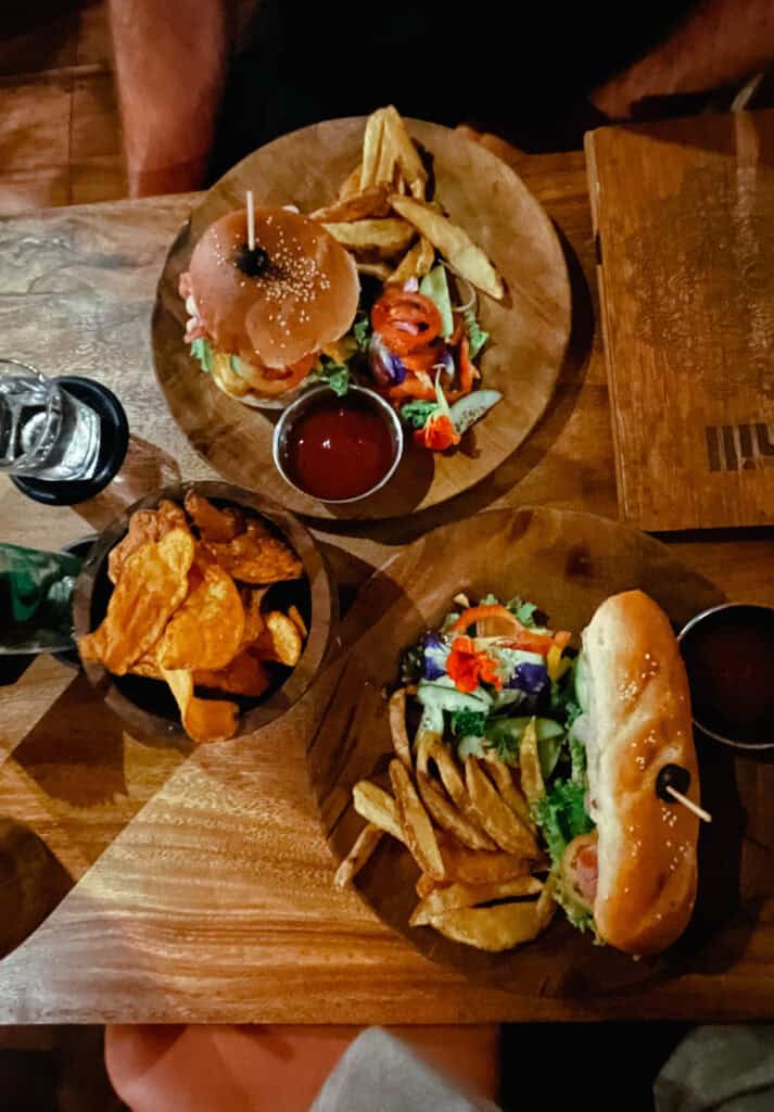 Overhead view of a wooden table with two gourmet burger plates, each served with fries, salad garnished with edible flowers, and a side of ketchup with sweet potato fries.