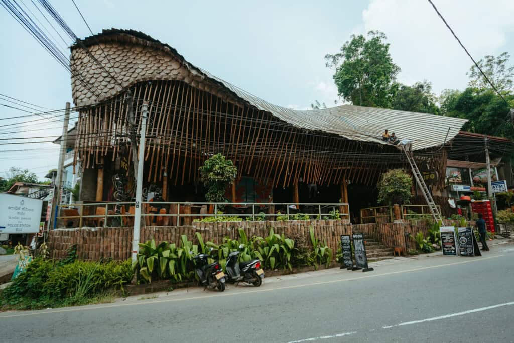Street view of a uniquely designed café in Ella, Sri Lanka, featuring a wavy roof made of bamboo and wood. The exterior is lined with greenery, rustic fencing, and chalkboard signs advertising menu items.