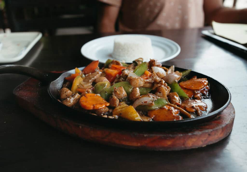 Close-up of a sizzling plate of stir-fried vegetables and chicken, including bell peppers, onions, carrots, and zucchini, served on a wooden board.