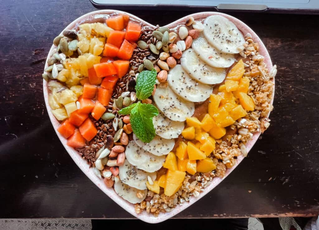 Overhead view of a vibrant smoothie bowl served in a heart-shaped bowl, topped with sliced bananas, diced mango, and other fruits.