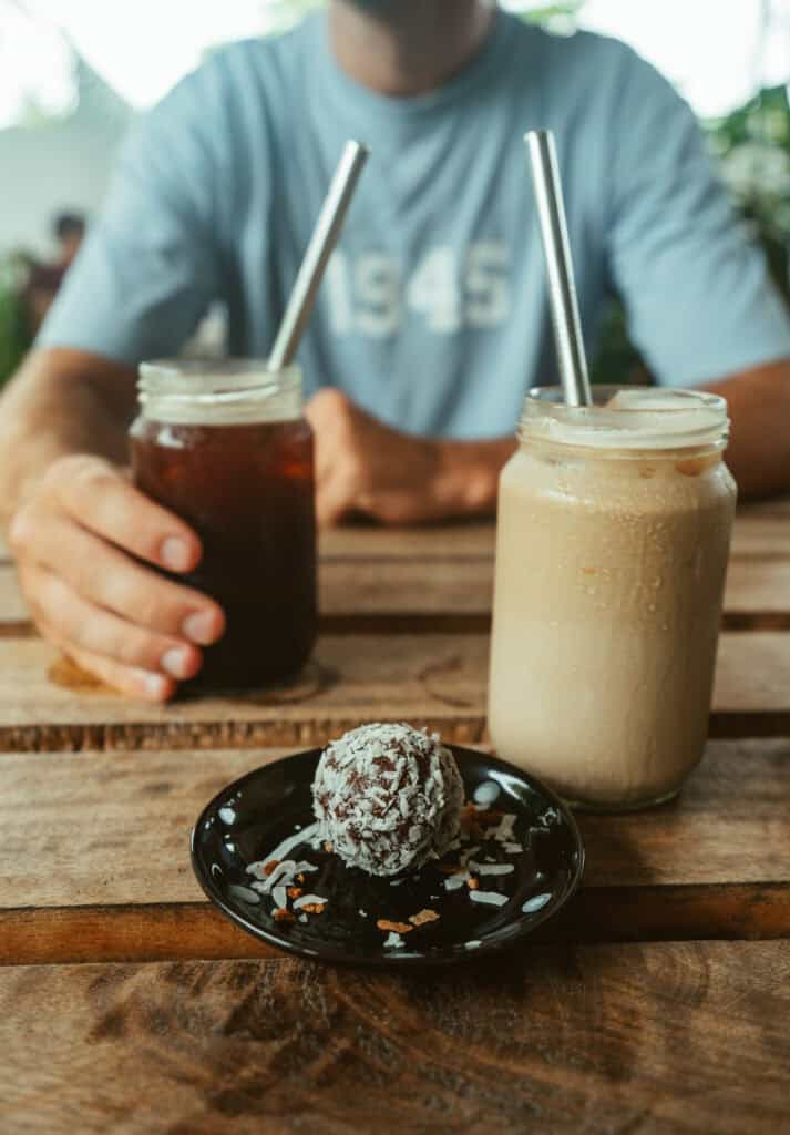Close-up of a chocolate energy ball coated with coconut flakes on a black plate, accompanied by two mason jars filled with iced coffee and a latte from a Cafe in Marissa, Sri lanka